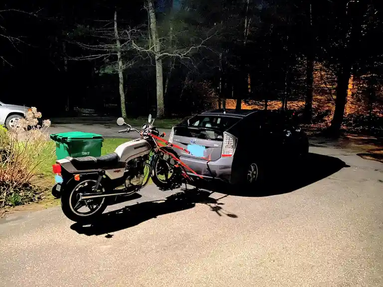 A silver Honda motorcycle is parked next to a gray Toyota Prius at night, with bright orange straps visibly securing the…