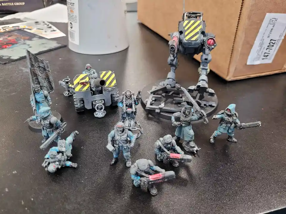 A collection of painted miniature sci-fi soldiers and a large robot stand on a dark tabletop, surrounded by hobby supplies…