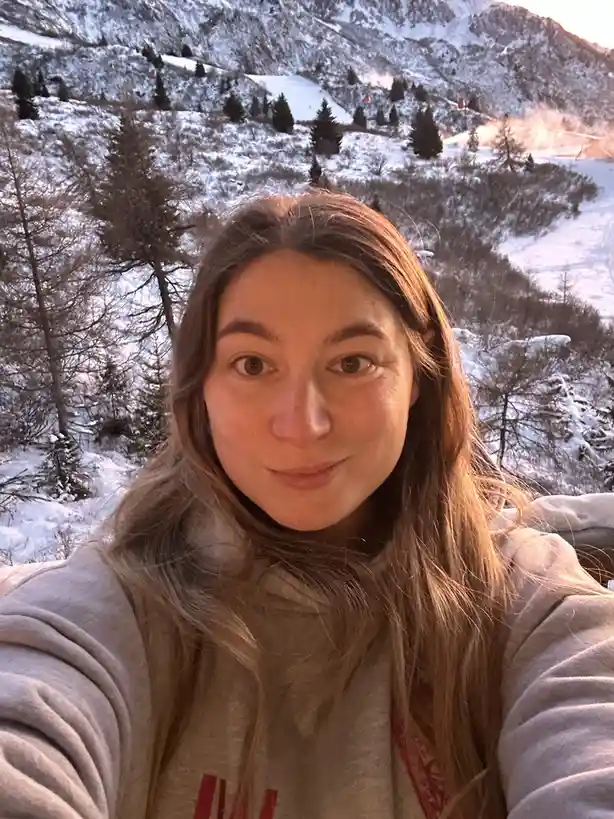 A selfie of a woman with long brown hair wearing a grey hoodie, taken outdoors in a snowy mountain landscape with evergreen…