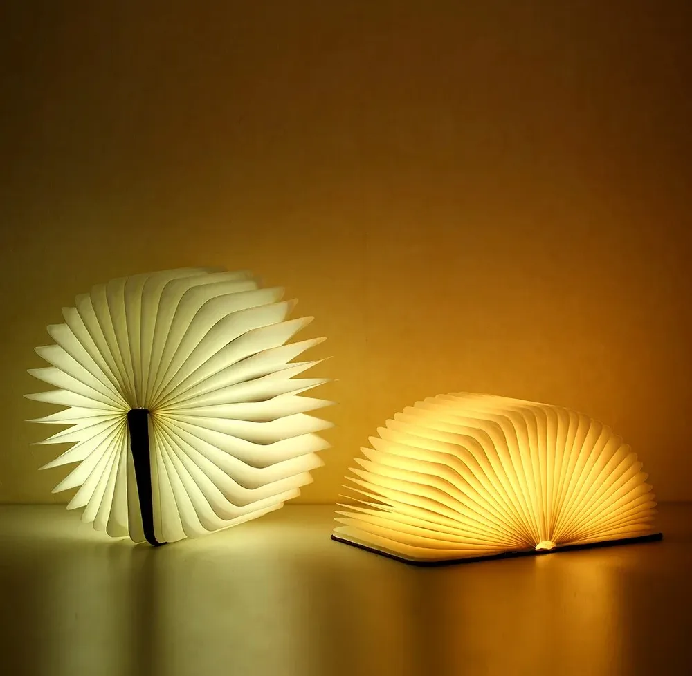 Two illuminated, fan-shaped book lamps sit on a surface against a warm-toned wall; one is upright and glowing white, the…