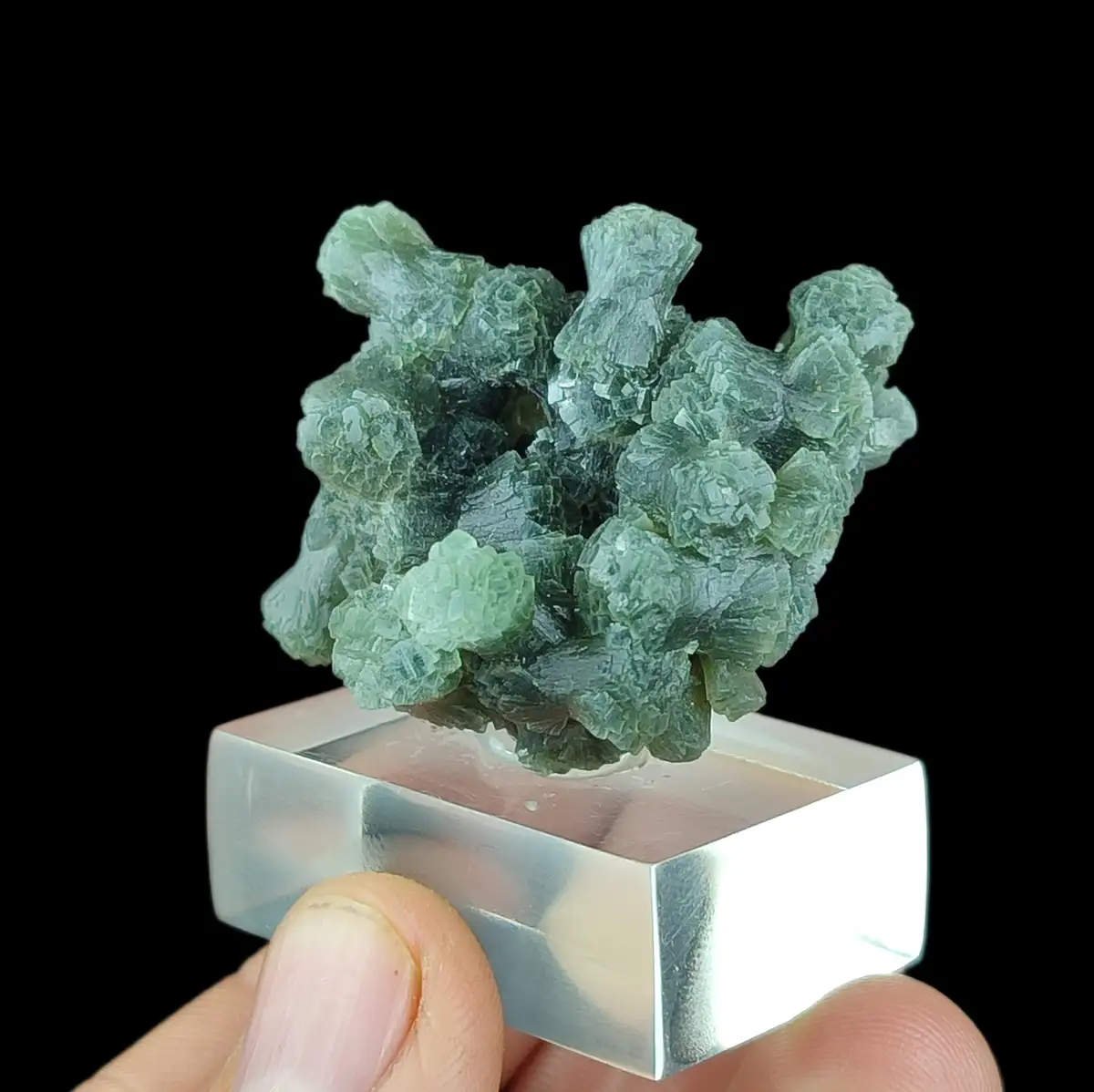 A hand holds a small, silver-colored square stand displaying a cluster of translucent, greenish-blue crystals that resemble…
