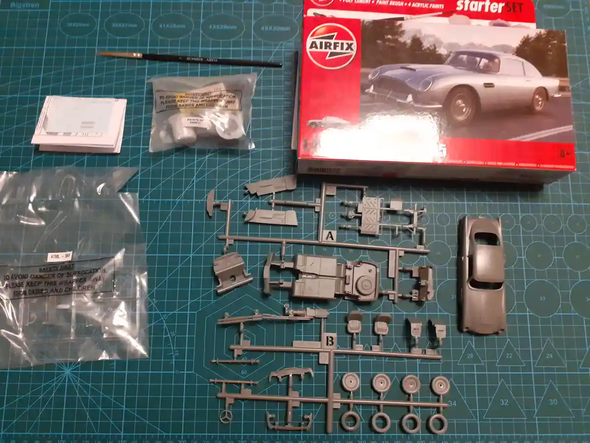 An Airfix 1:4 scale model car starter kit is laid out on a green cutting mat, showing plastic parts, instructions, paint…