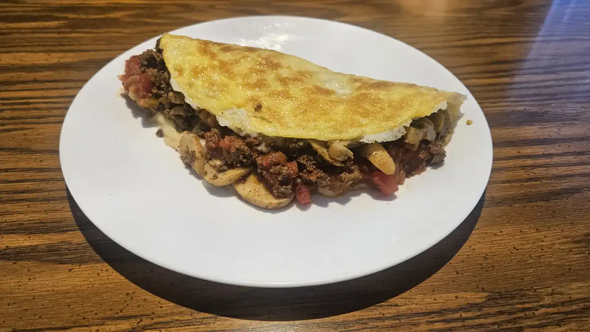 A close-up photo shows a folded, golden-brown omelette on a white plate, revealing a filling of ground meat, diced tomatoes…