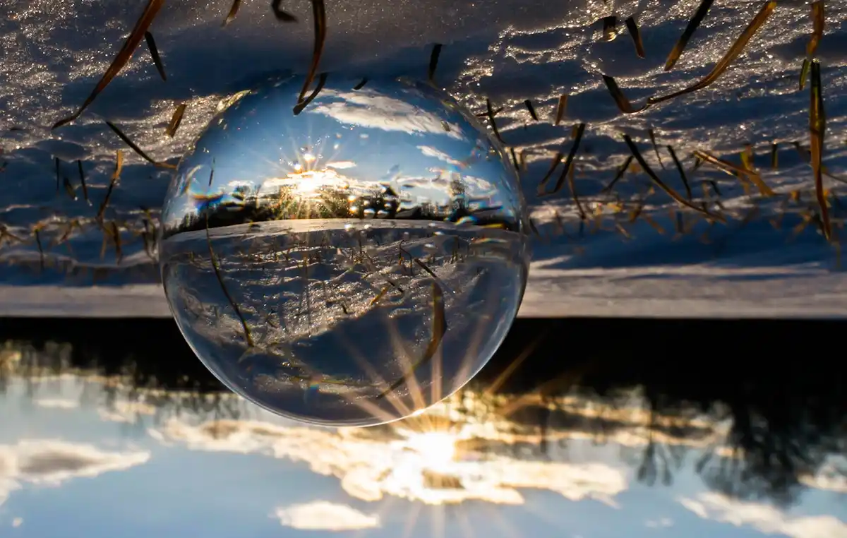 A clear glass sphere rests on snowy ground, reflecting a bright sun and sky with lens flare, surrounded by dry grass stalks…
