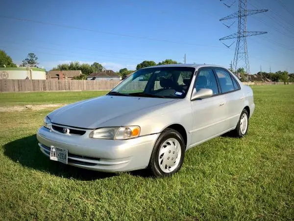 A silver Toyota Camry sedan is parked on a grassy field under a clear blue sky, with power lines and a fence visible in the…