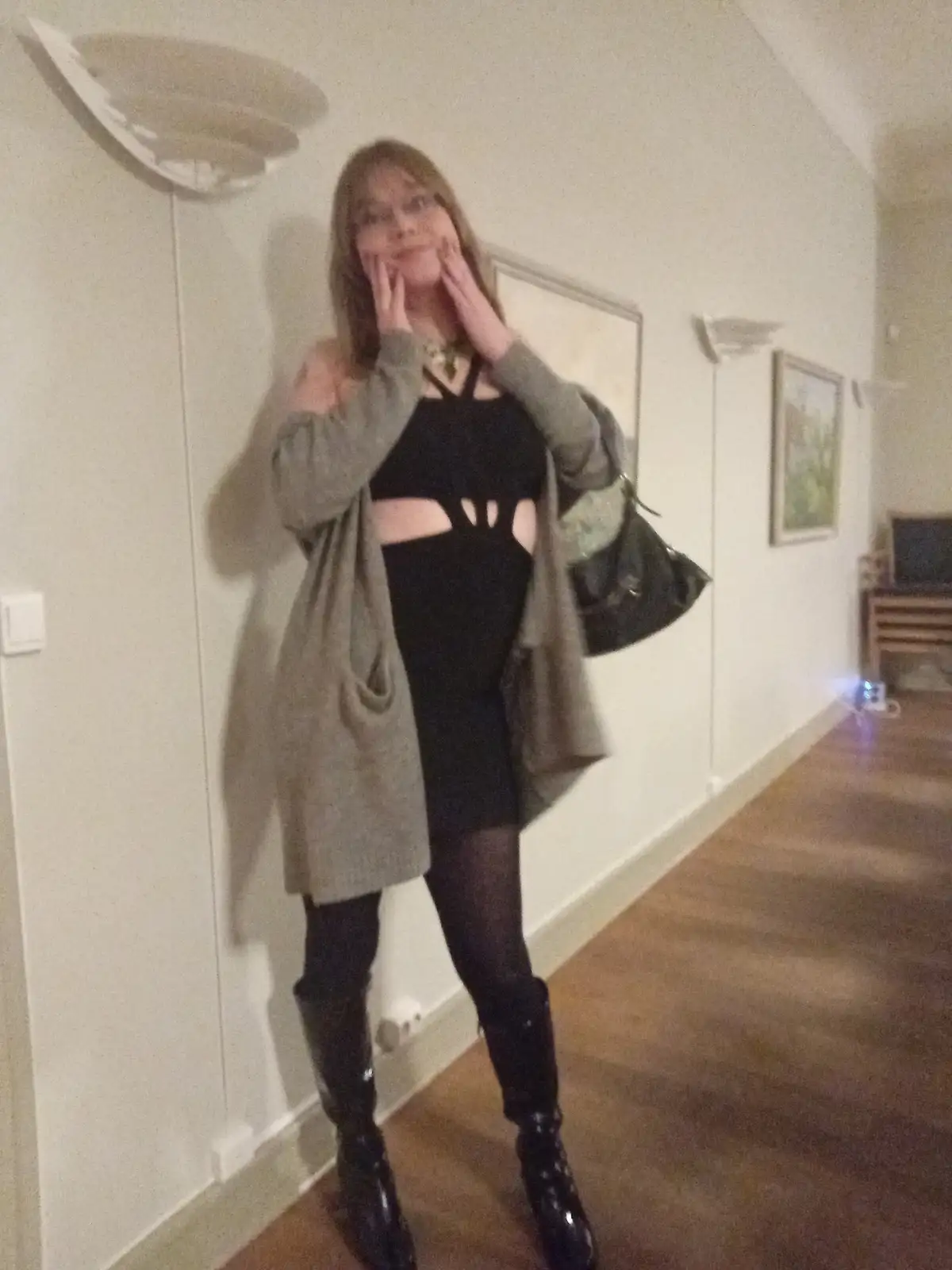 A person with glasses and long hair stands in a hallway wearing a black cutout dress, a grey open cardigan, black tights…