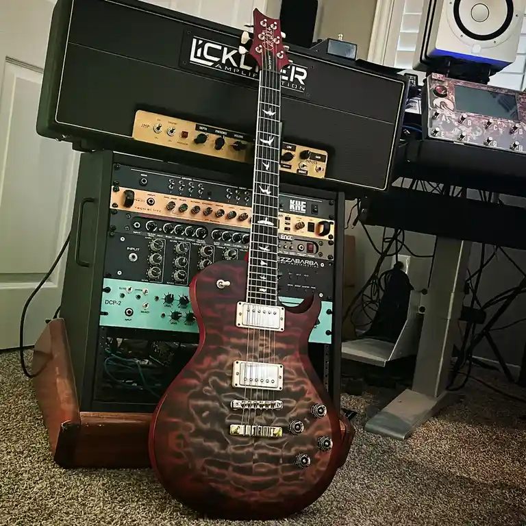A red and black quilted finish electric guitar leans against a tall stack of professional audio equipment including a…