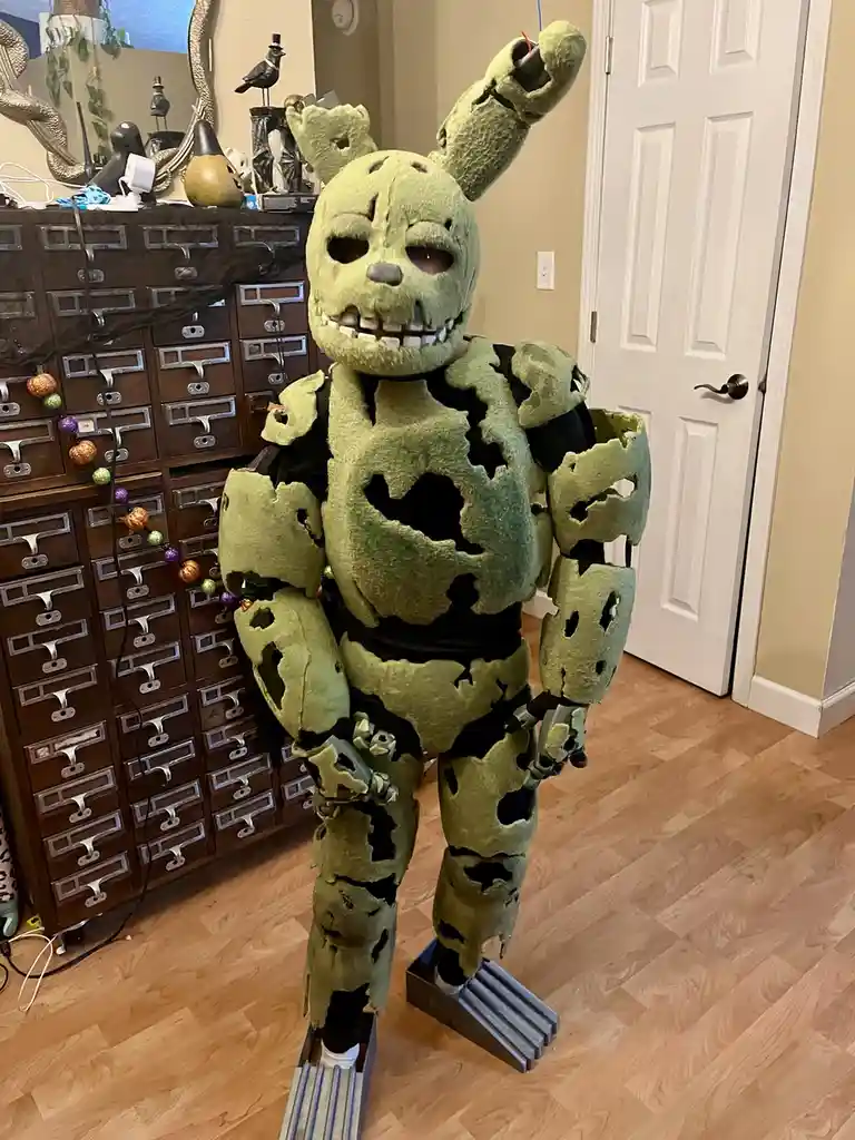 A person in a tattered, green, animatronic bunny costume with exposed mechanical parts and a skull-like face stands indoors…