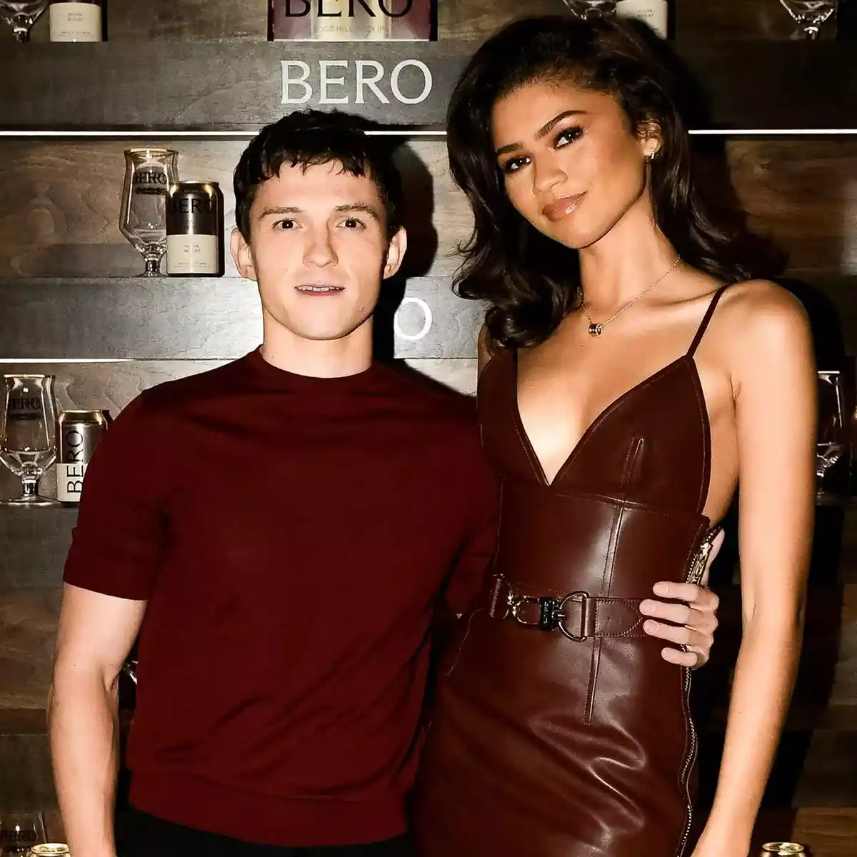 A man and a woman pose together in front of a bar with 'BERO' branding on shelves and glassware. The man wears a plain…
