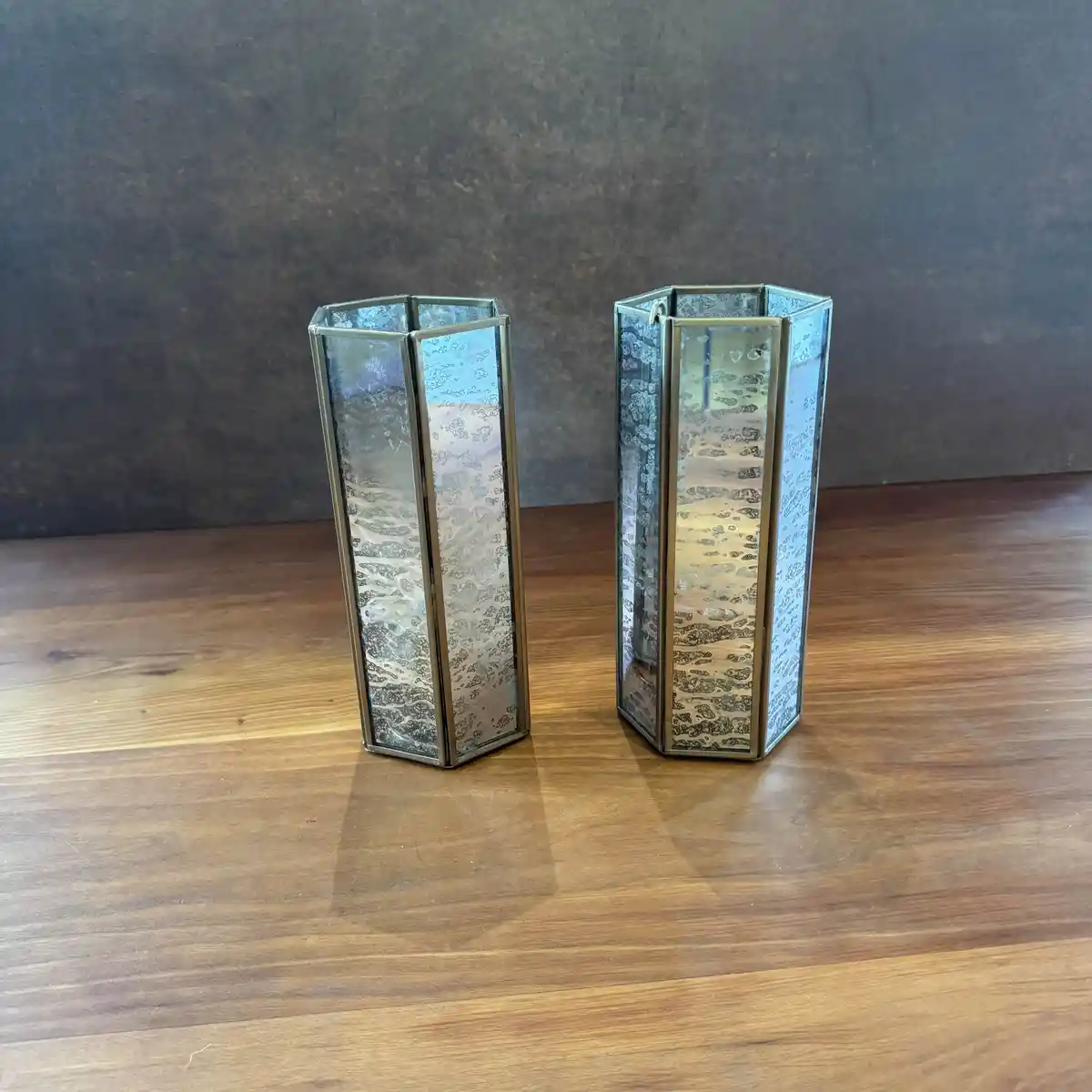 Two hexagonal glass candle holders with textured, iridescent glass panels and brass-colored metal frames sit side by side on…