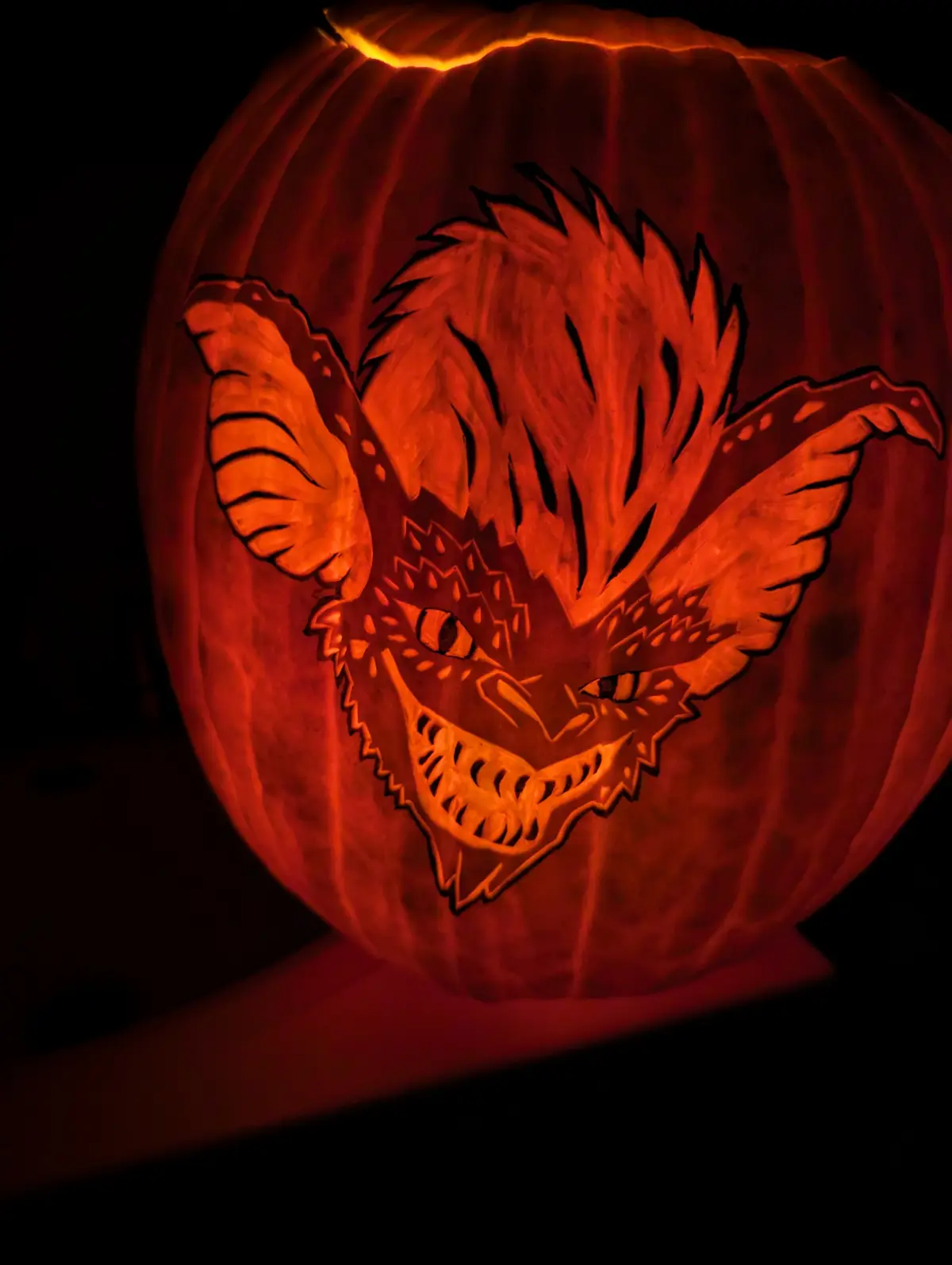 A close-up of a carved Halloween pumpkin illuminated from within, featuring a detailed demonic face with bat-like wings…