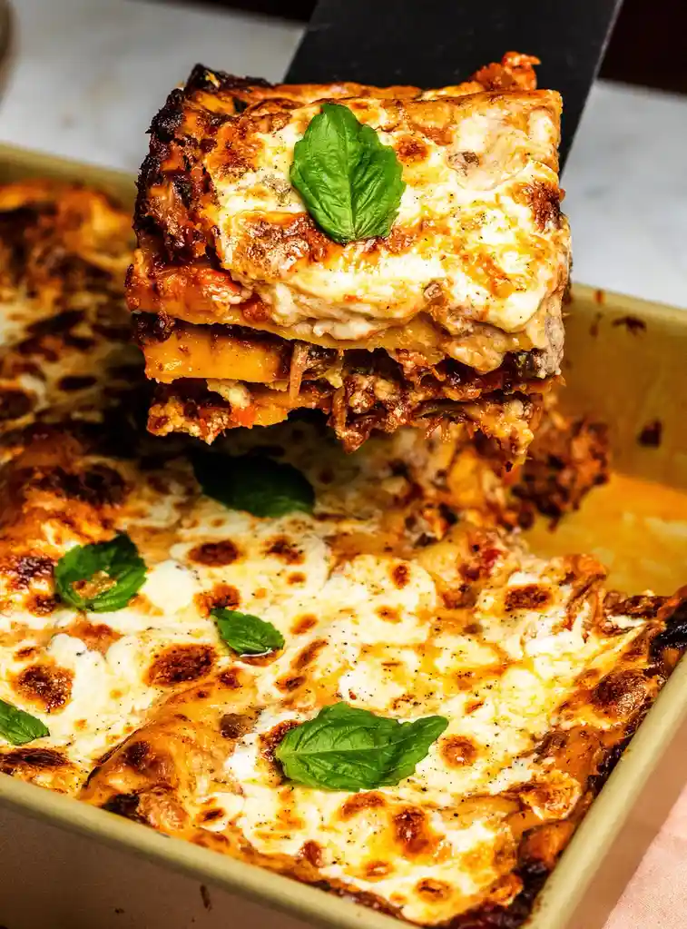 A close-up photo shows a black spatula lifting a perfectly baked, cheesy lasagna slice from a baking dish. The slice reveals…