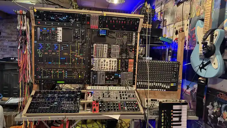 A densely packed, custom-built modular synthesizer setup with numerous black and colored modules, knobs, and illuminated…