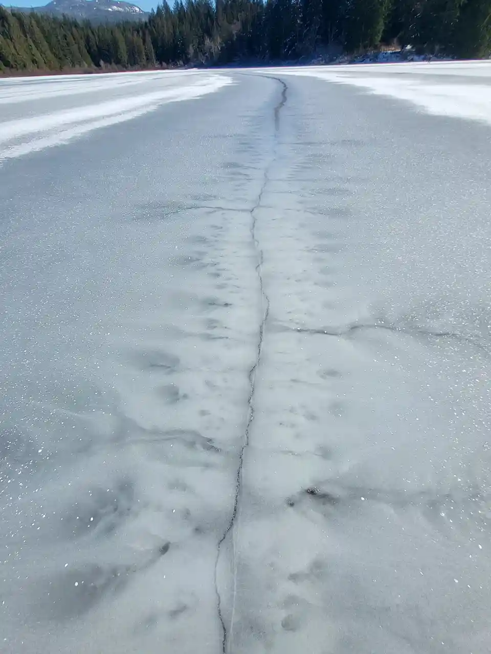 A wide, frozen lake covered in snow and ice, with a prominent, winding crack running down the center. In the background, a…