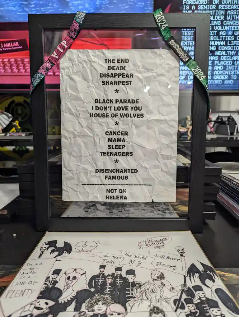 A crumpled paper list of song titles is displayed inside a black picture frame, secured with two event lanyards (one pink…