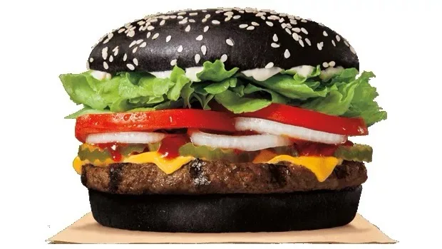 a black sesame seed bun burger with visible layers of lettuce, tomato, onion, pickles, melted cheese, and ketchup on a white…