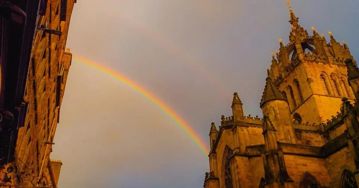 A low-angle photo shows a vibrant rainbow arcing across a twilight sky between two ornate, gothic-style stone buildings. The…