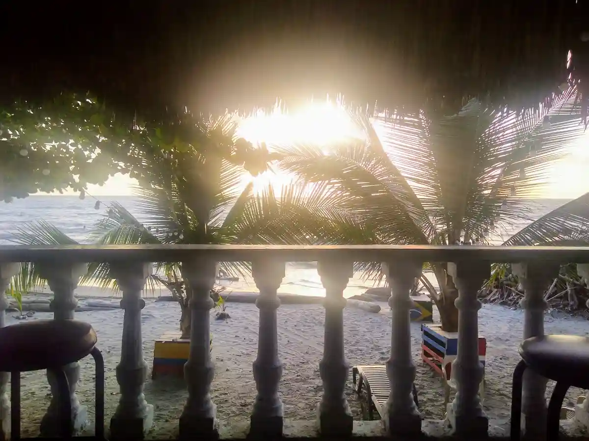 A serene tropical beach scene viewed from a thatched-roof balcony with ornate stone balusters, palm fronds silhouetted…