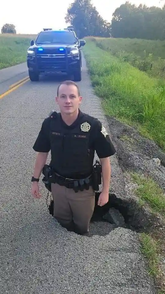 A police officer in uniform stands with his lower body stuck in a large hole in a rural road, looking directly at the camera…