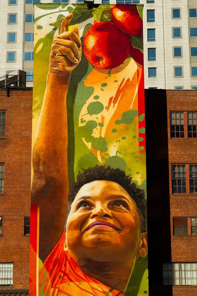 A large mural on a brick building depicts a smiling Black woman with short curly hair, wearing an orange top, looking upward…