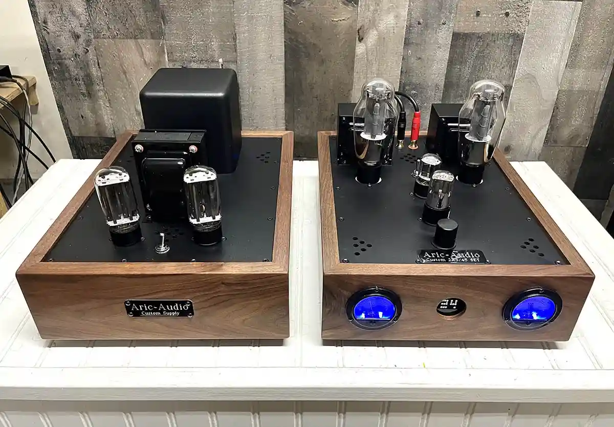 Two wooden-cased audio amplifiers with visible vacuum tubes and glowing blue dials sit side by side on a white surface. The…