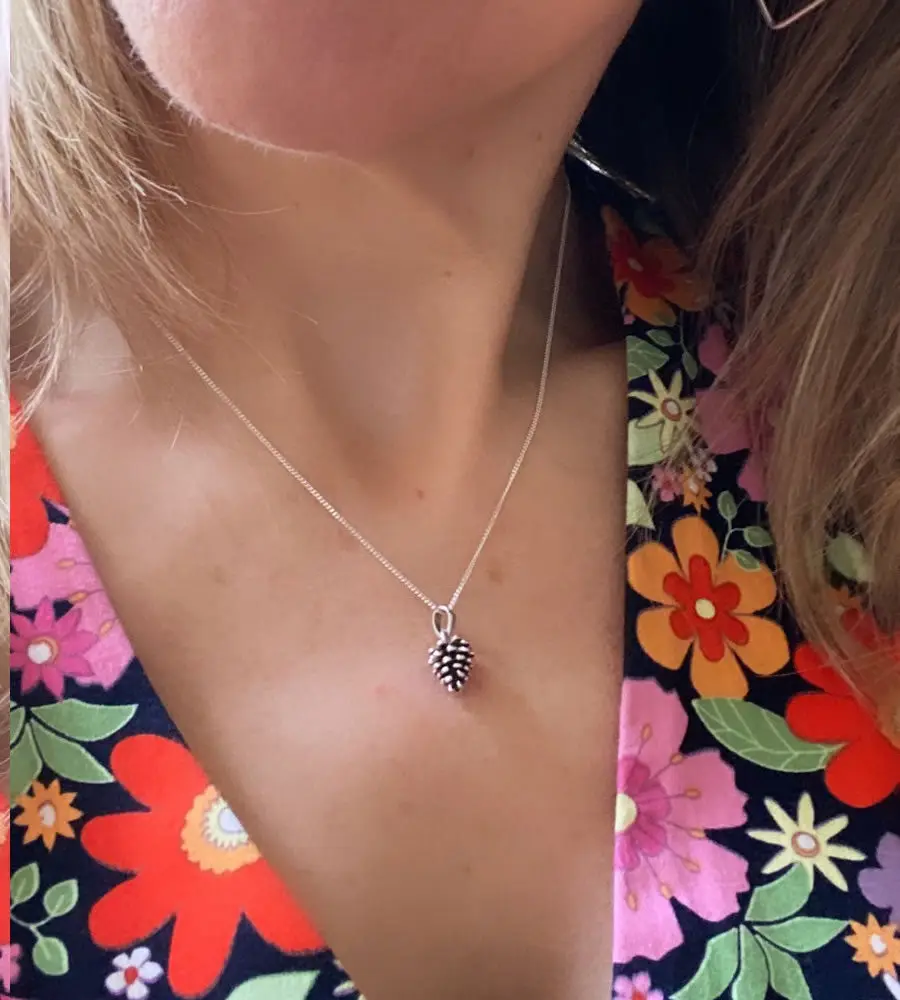 A close-up photo shows a person wearing a silver chain necklace with a small pinecone-shaped pendant centered on their…
