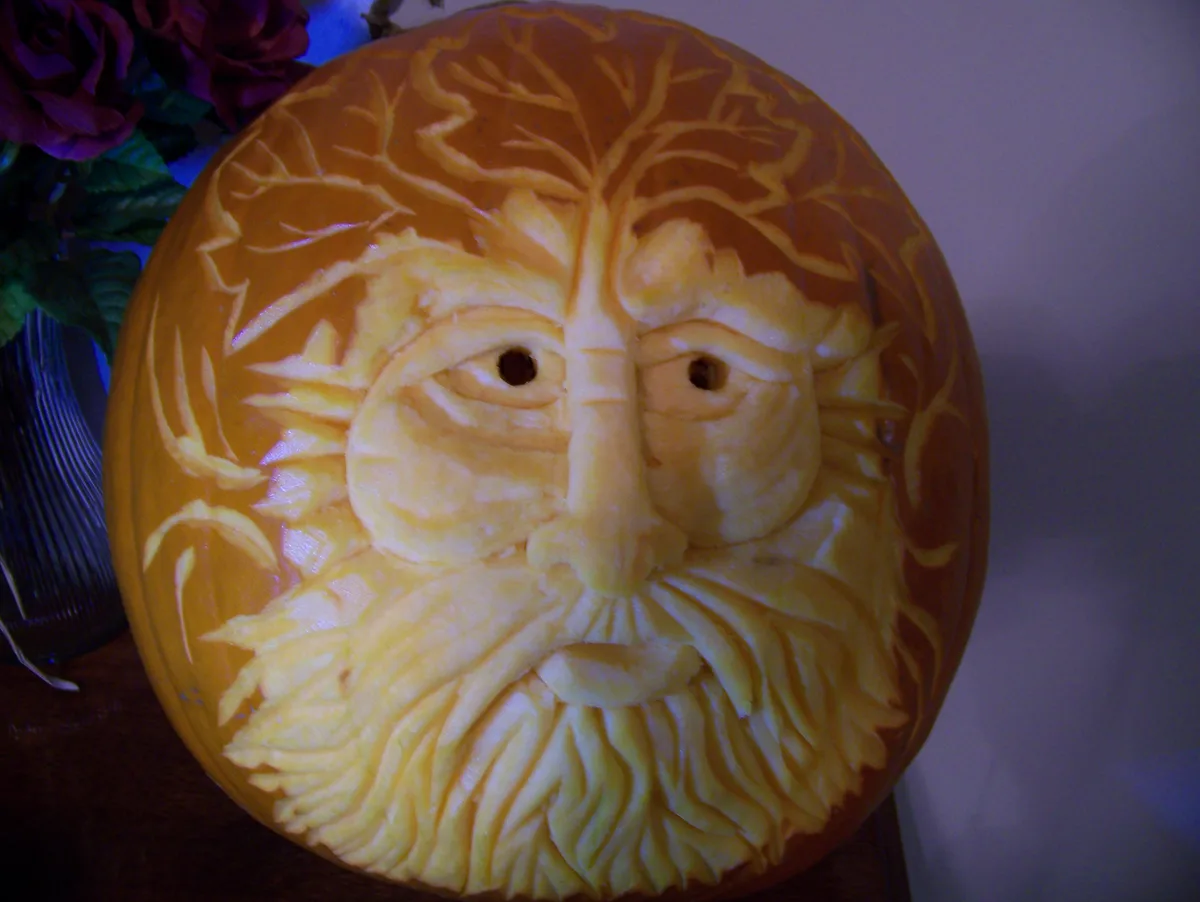 A close-up of a meticulously carved pumpkin featuring a face with tree-like hair and beard, resembling a wise old man or…