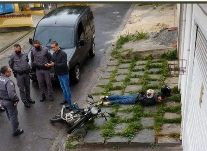 A man lies unconscious on a grassy sidewalk next to a fallen motorcycle, while several police officers stand nearby…