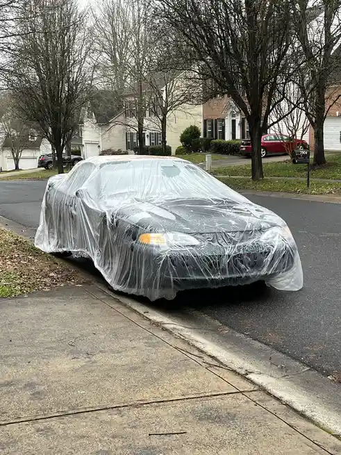 A dark-colored car is parked on a residential street, completely covered by a transparent plastic sheet that is visibly…
