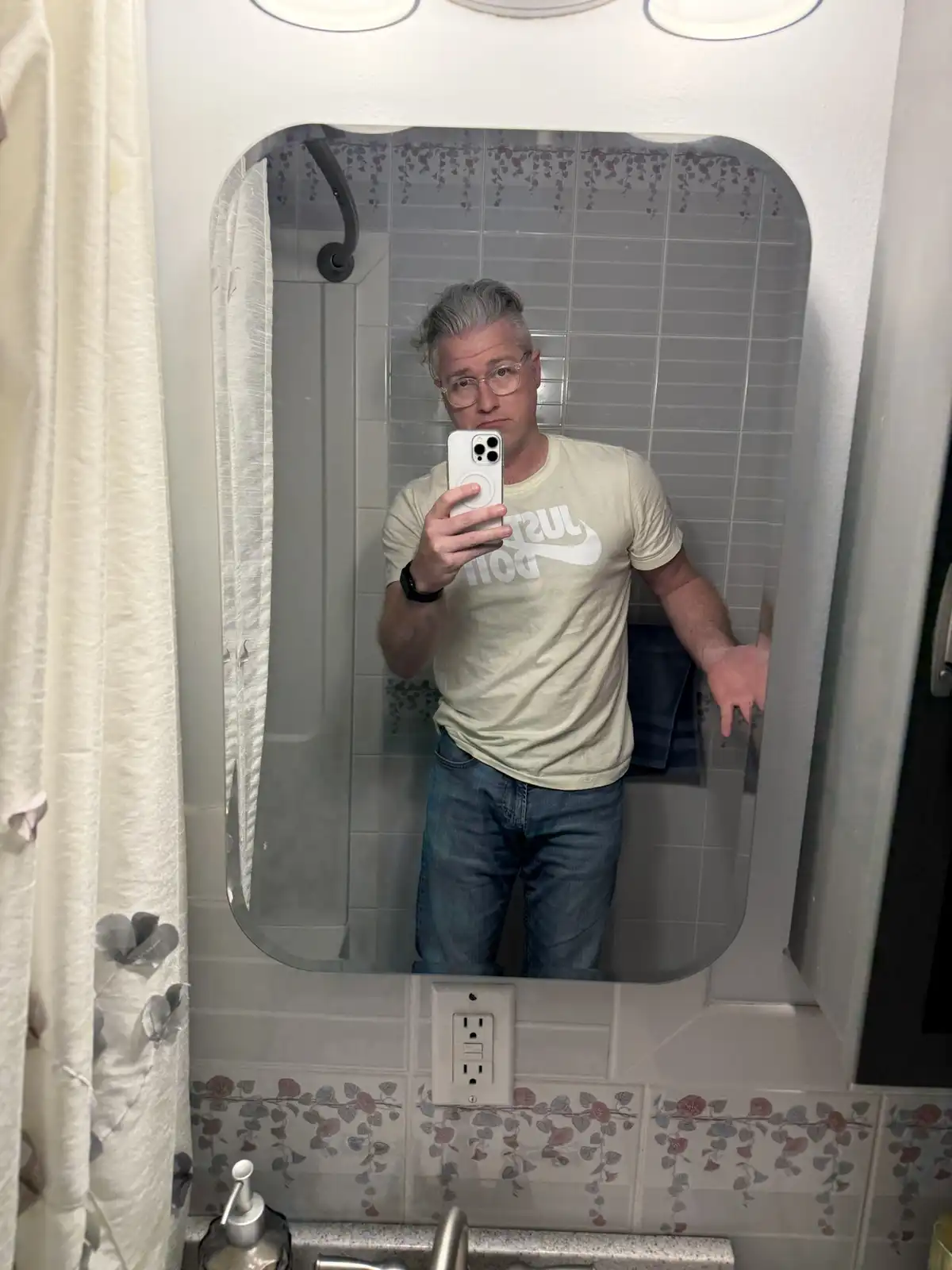 A man with gray hair and glasses takes a mirror selfie in a bathroom, wearing a beige t-shirt with the reversed text 'just…