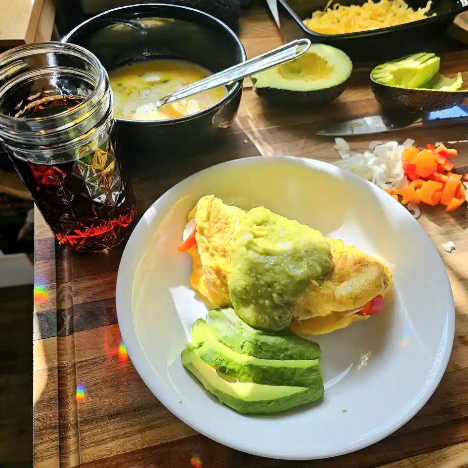 A plate of an omelet topped with a large dollop of mashed avocado, with sliced avocado on the side, surrounded by…