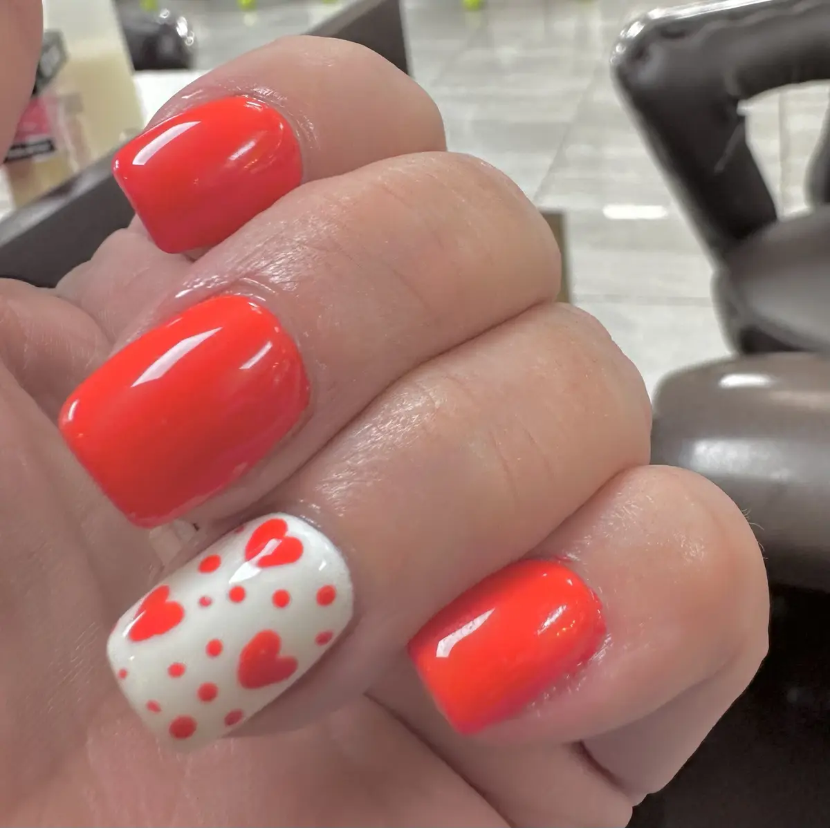 A close-up photo shows a hand with glossy, bright coral-pink fingernails, except for one finger which has a white base with…
