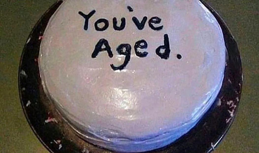 A close-up of a round cake with white frosting, sitting on a dark plate. The cake has the phrase 'You've Aged.' written on…
