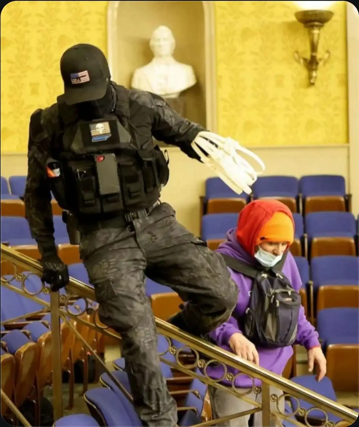 A person in full tactical gear, including a black mask and vest with skull patches, is descending stairs while holding a…