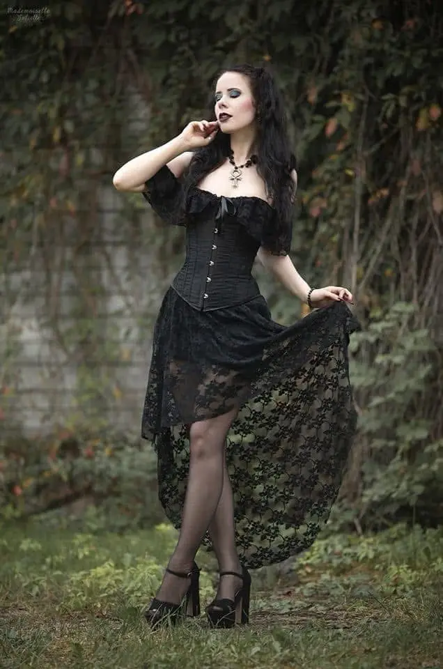 A woman with long dark hair, pale makeup, and dark lipstick poses outdoors in a black corset dress with lace overlay and…