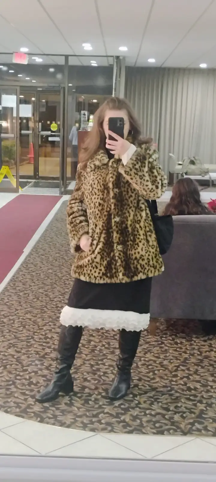 A person takes a mirror selfie in a hotel lobby wearing a leopard-print faux fur coat, a black skirt with a white ruffled…