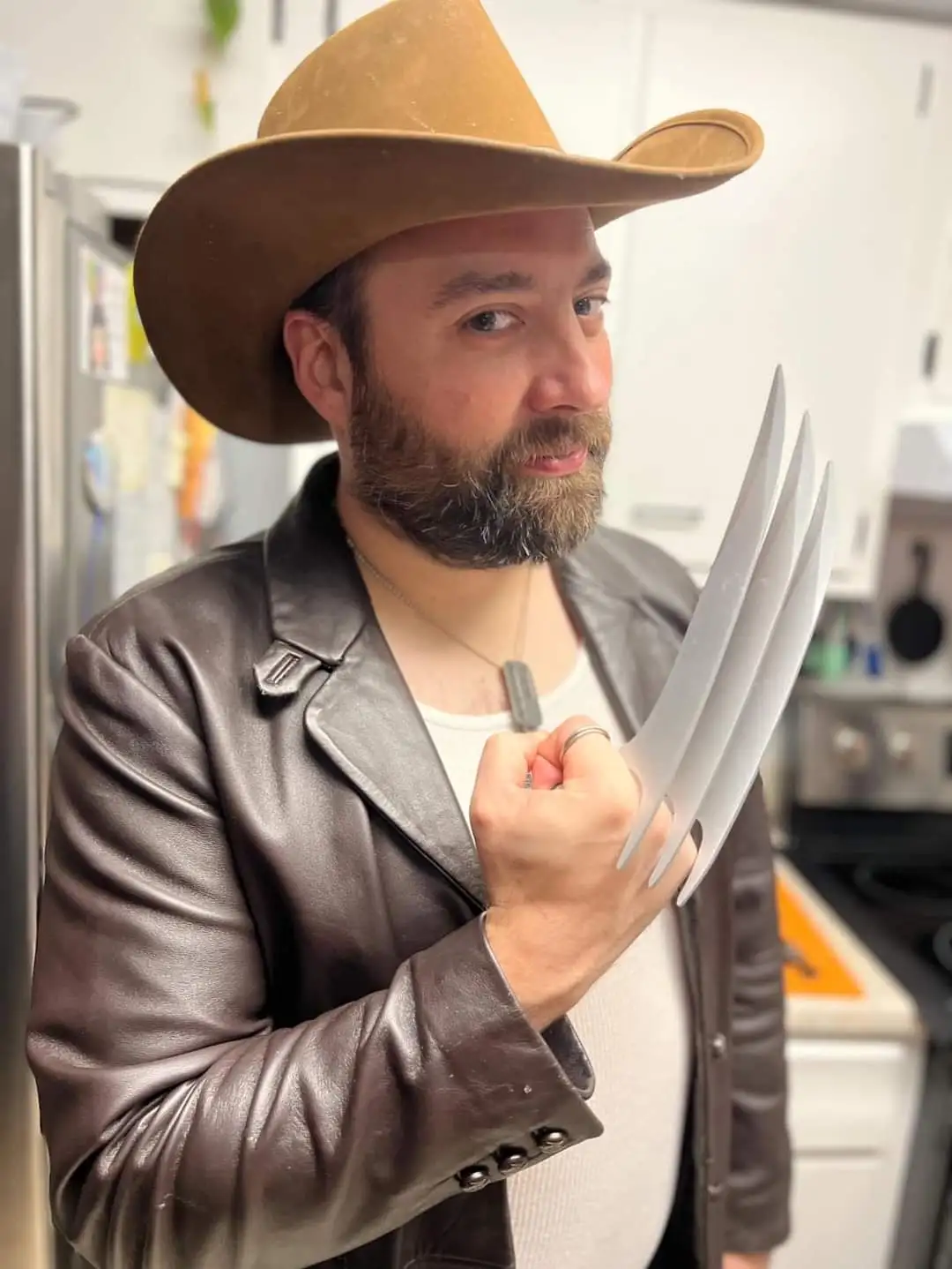 A man with a beard wearing a brown cowboy hat, a brown leather jacket over a white shirt, and a dog tag necklace, stands in…
