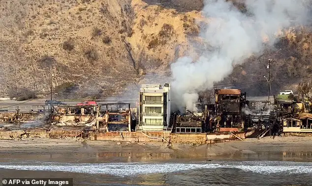 a photograph shows a beach with ocean waves in the foreground and a row of buildings behind it, most of which are charred…