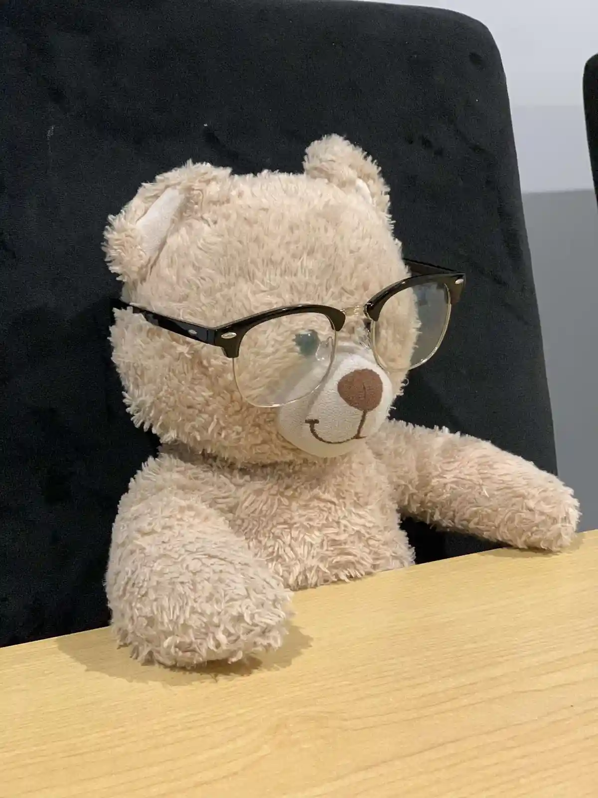 A beige plush teddy bear wearing black-rimmed glasses sits upright in a black office chair with its arms resting on a light…