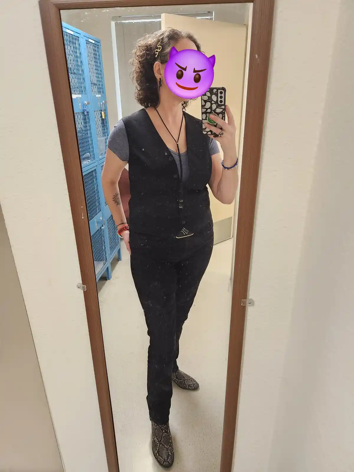 A person takes a mirror selfie in a fitting room, wearing a black vest over a grey shirt, black pants, and snakeskin boots…