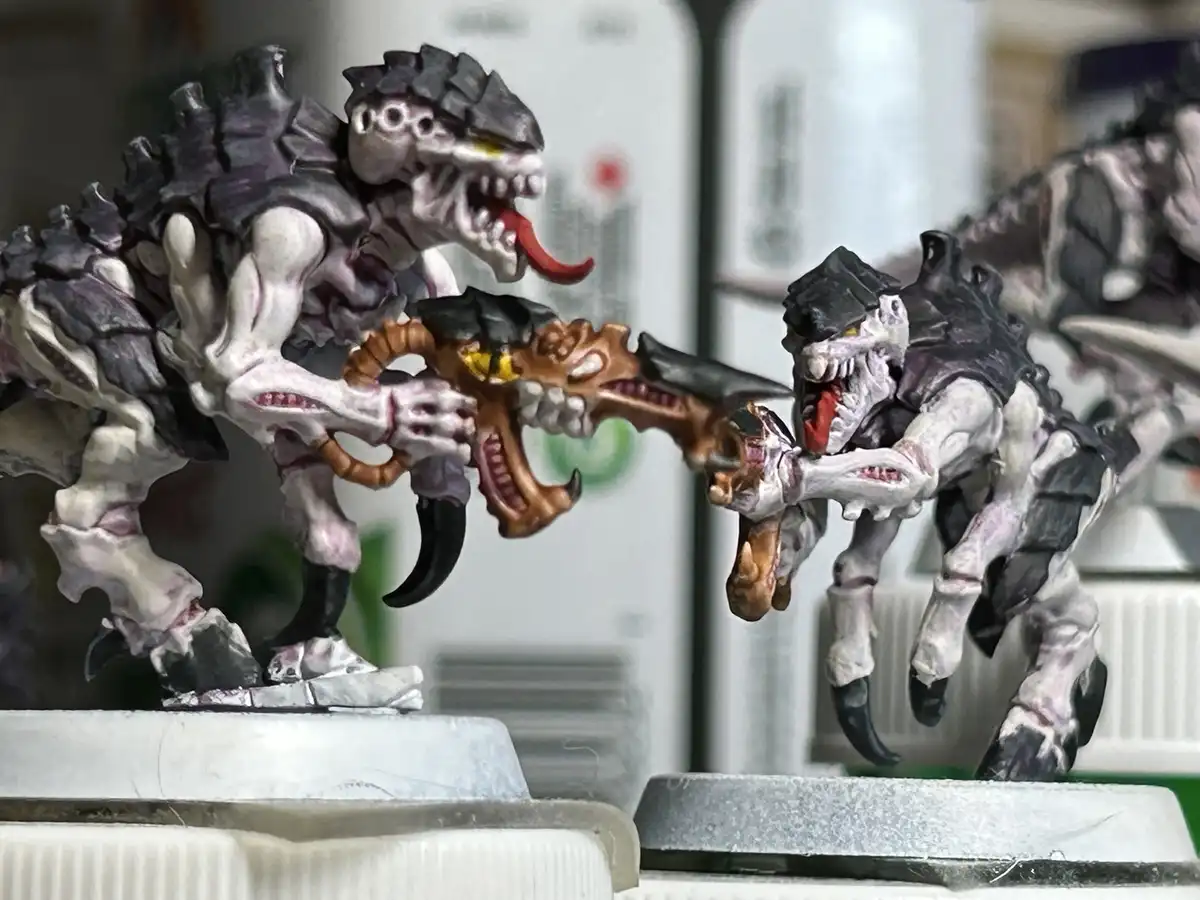 Two painted fantasy miniatures, resembling grotesque, skeletal, reptilian creatures with sharp teeth and claws, are locked…