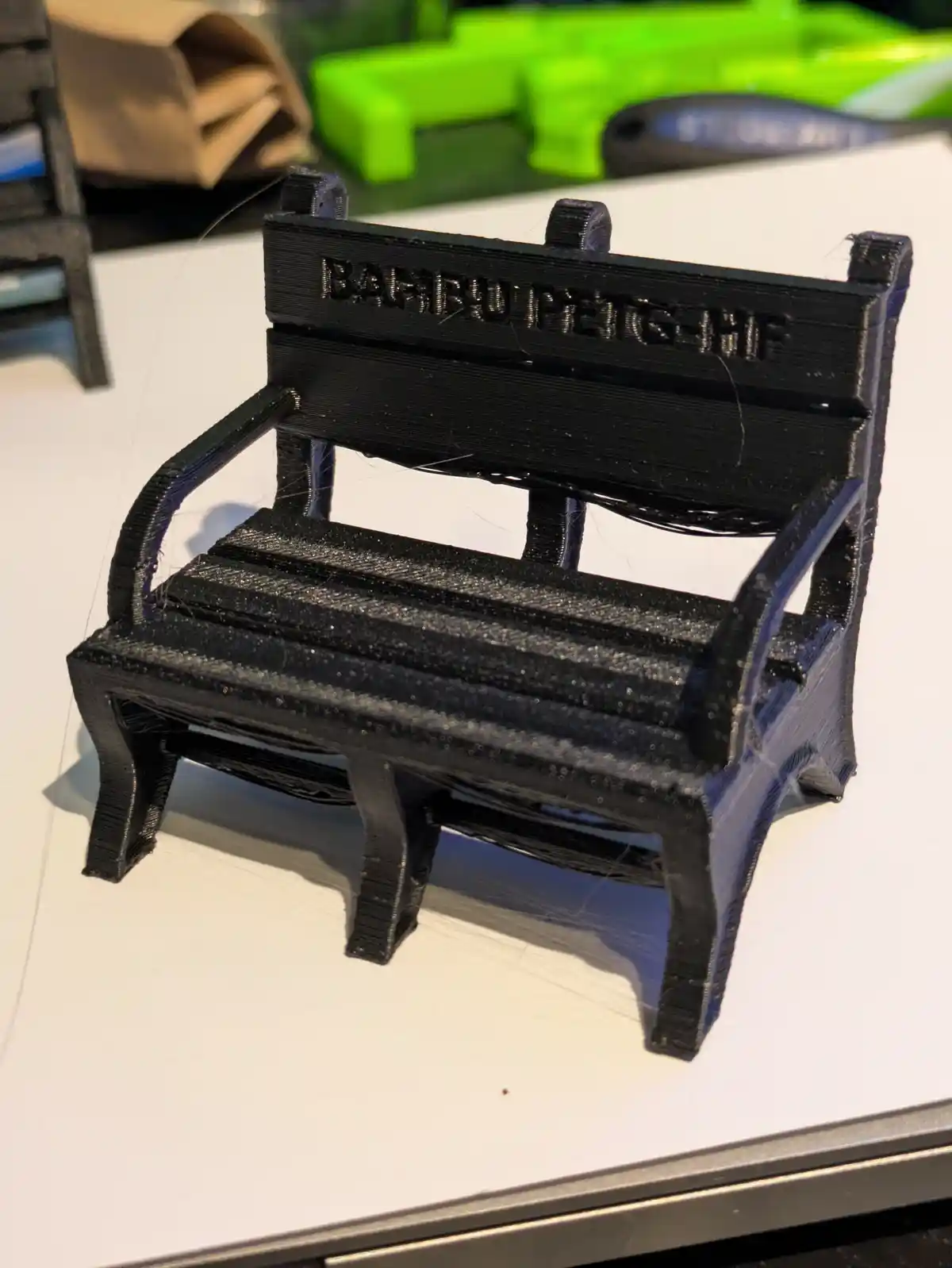 A small, black 3D-printed miniature bench sits on a white surface, showing visible layer lines from the printing process…