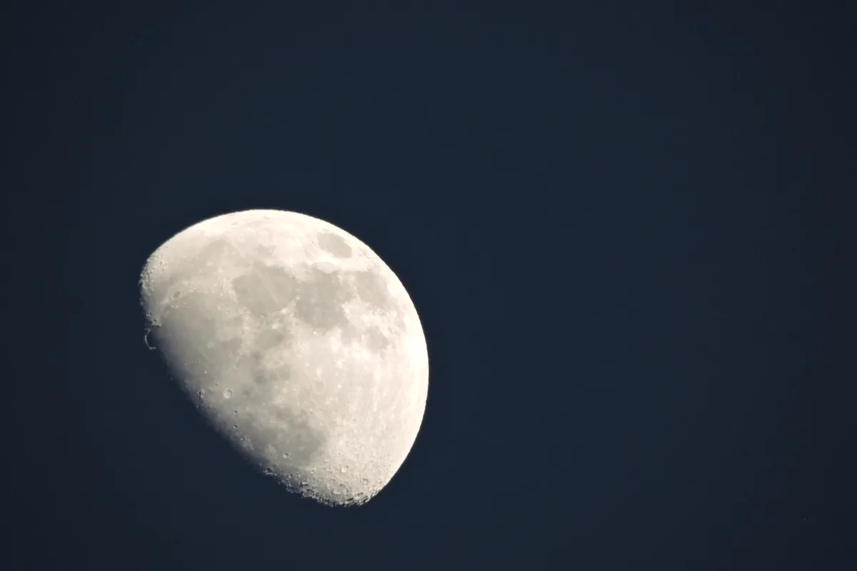 A close-up photograph of the moon against a dark blue sky, showing its cratered surface and illuminated crescent. The image…