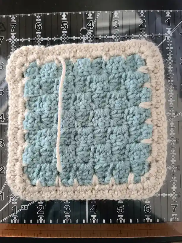 A handmade crocheted square with a beige border and light blue center sits on a black cutting mat with grid lines and…
