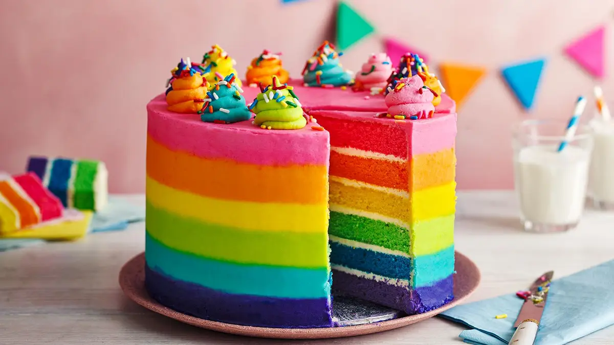 A vibrant, multi-layered rainbow cake with a slice cut out, revealing its colorful interior. The top is decorated with…