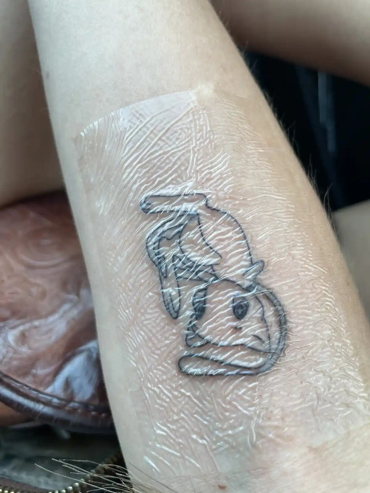 A close-up photo shows a freshly tattooed design on a person's forearm, covered with a clear plastic wrap. The tattoo is a…