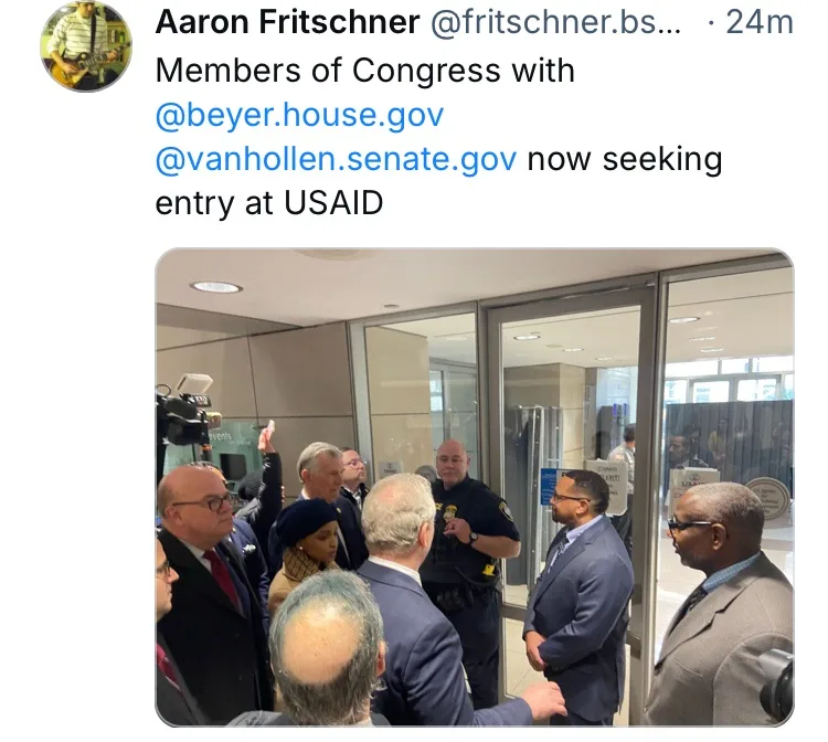 A group of men in suits, including recognizable congressmen, are gathered outside a glass door at USAID, appearing to be…