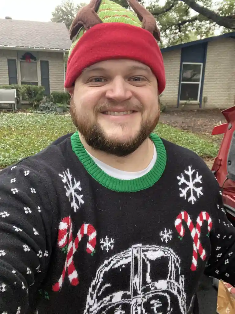 A man with a beard smiles at the camera while wearing a red beanie with brown reindeer antlers and a black Christmas sweater…