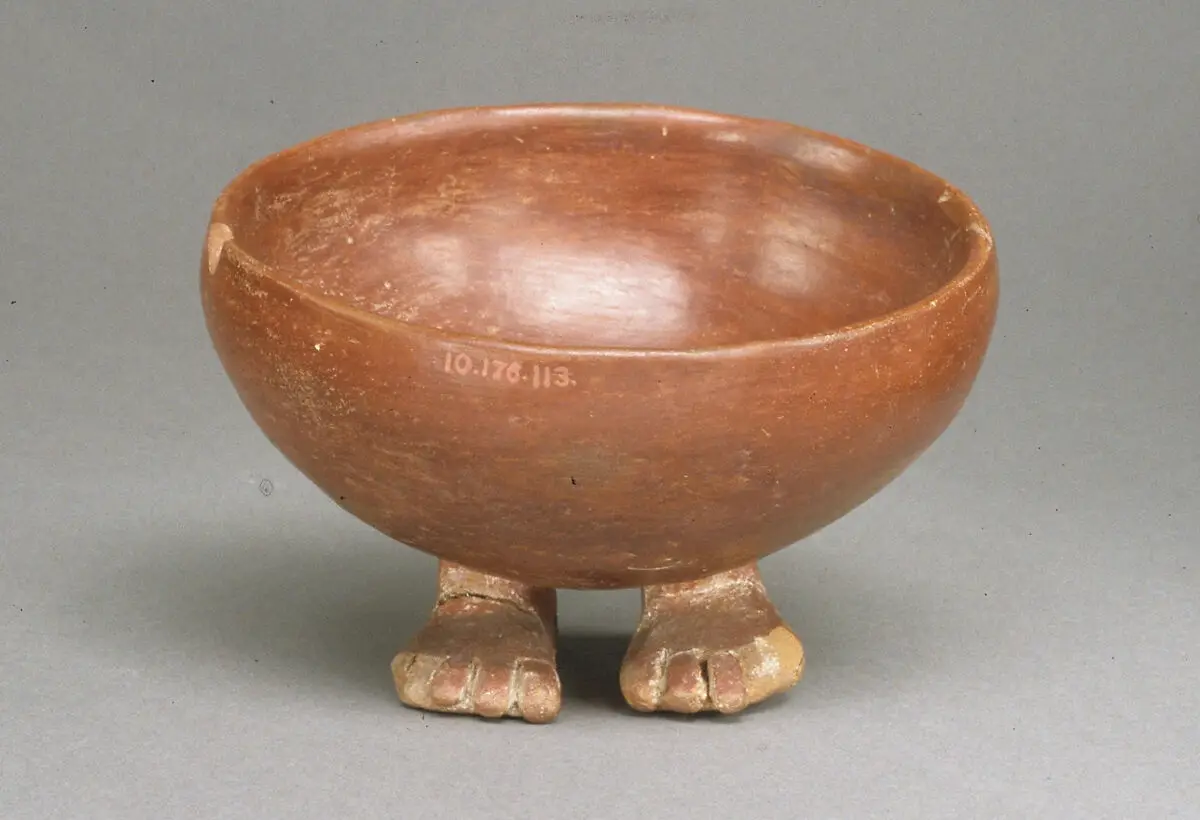 a brown ceramic bowl with two carved feet at its base, standing on a gray surface. The bowl has a museum accession number…