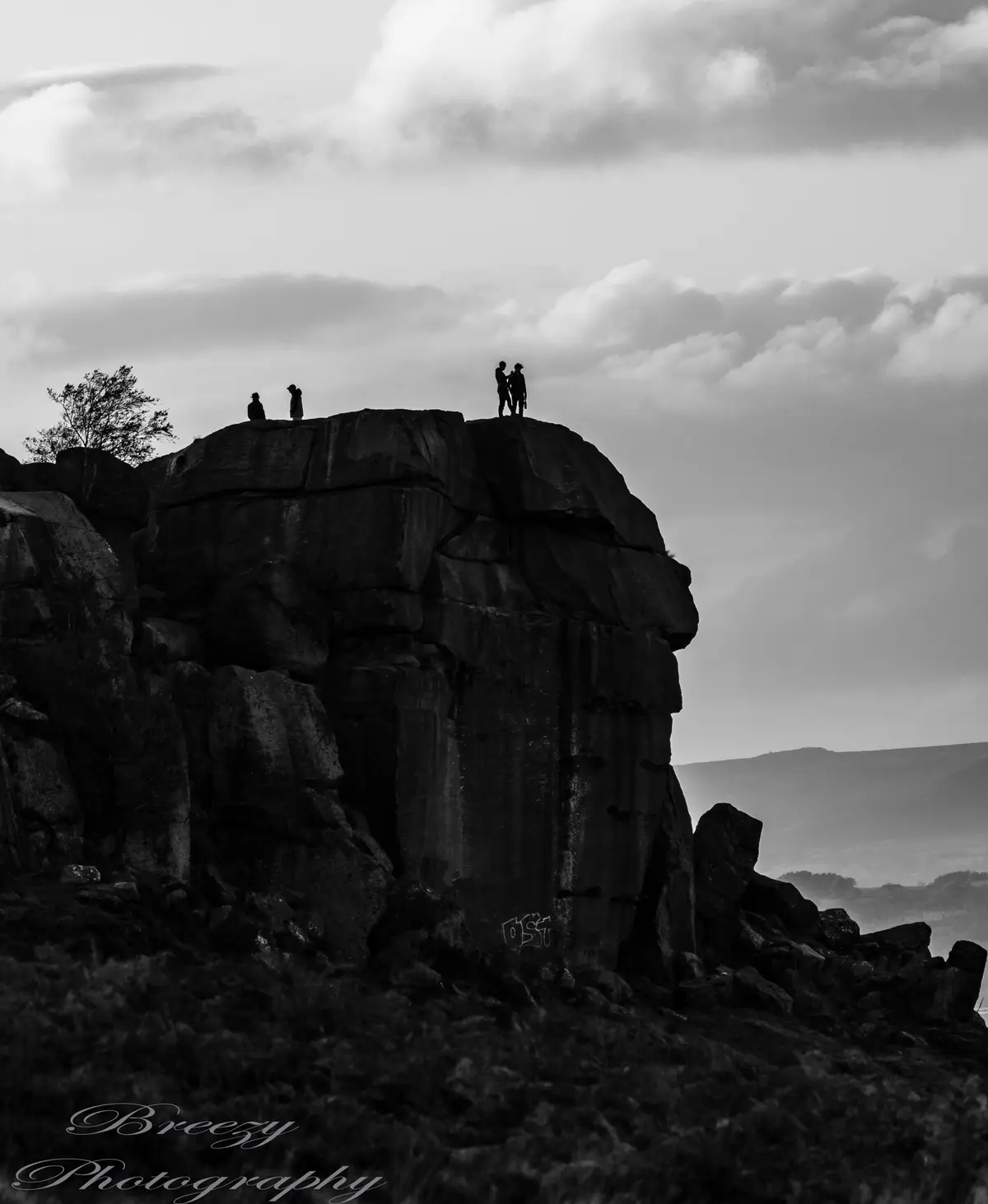 A black-and-white photo shows four silhouetted people standing atop a large, rugged rock formation under a cloudy sky. Two…